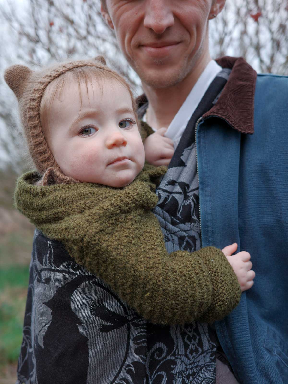 Baby in a knitted outfit with bear ears held in a Bairn carrier by an adult outdoors.