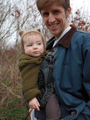 Man holding a baby in a Bairn carrier wearing a green outfit with a hood in an outdoor setting.