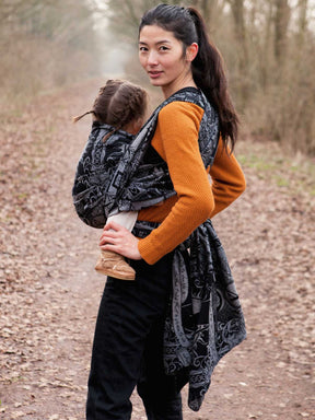 Woman carrying a child in a baby wrap outdoors on a path with trees in the background