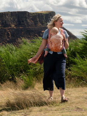 A mum carrying a baby in a colourful woven wrap with Arthur's seat in the background.