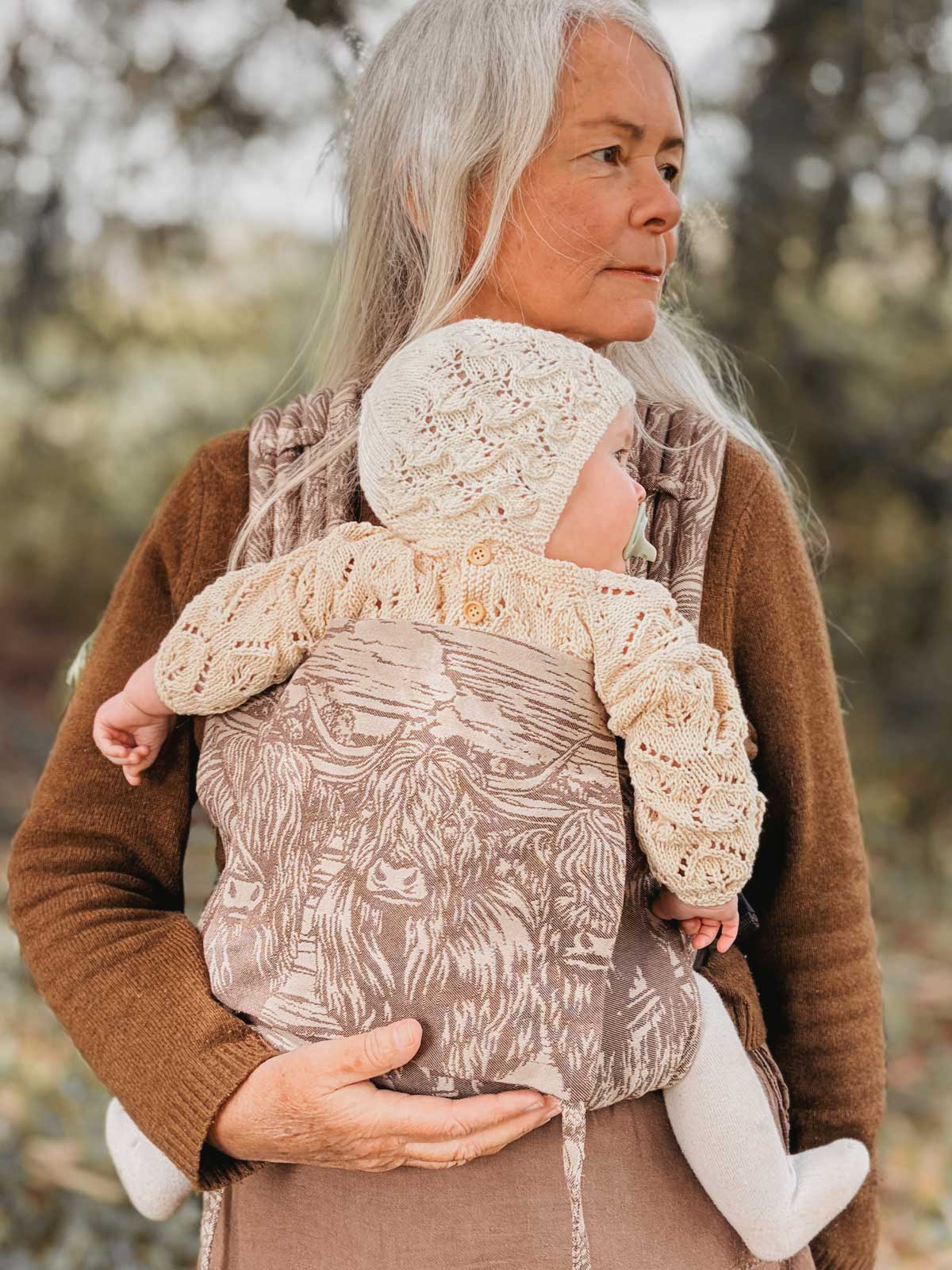 Woman holding a baby in a carrier with a highland cow pattern outdoors