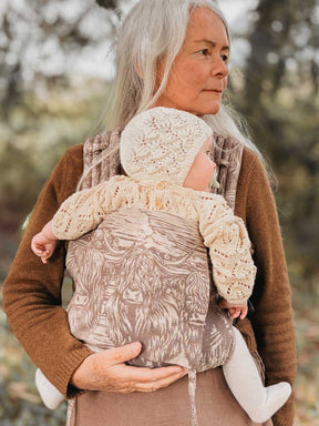 Woman holding a baby in a carrier with a highland cow pattern outdoors