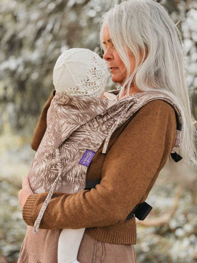 Woman carrying a child in a patterned carrier outdoors with snow-covered trees in the background