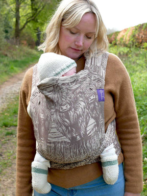 Woman carrying a baby in a baby carrier with a highland cow design in a rural setting.
