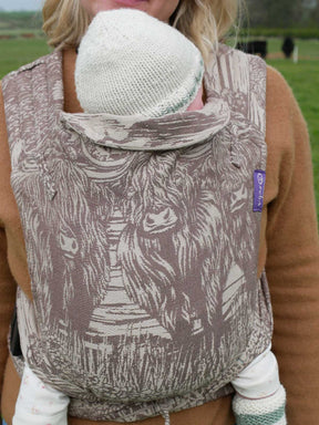 A baby in a baby carrier with a highland cow design in a rural setting.