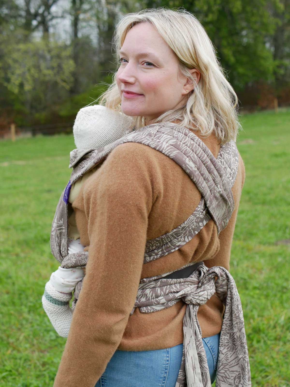 Woman carrying a baby in a baby carrier with a highland cow design in a rural setting.