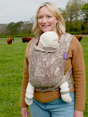 Woman carrying a baby in a sling outdoors with cows in the background