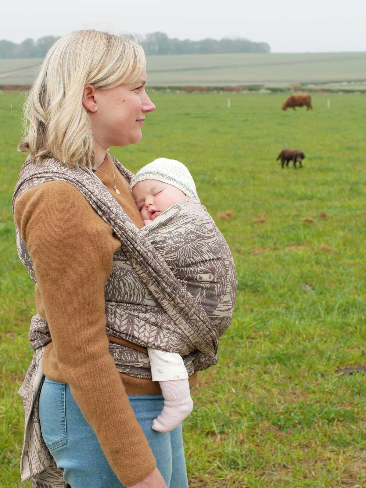 Woman carrying a baby in a sling outdoors in a field with cows in the background