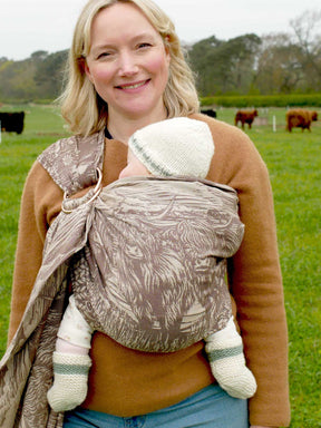 Woman carrying a baby in a sling outdoors with cows in the background