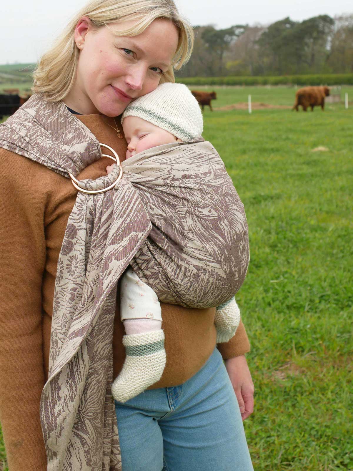 Woman holding a baby in a sling outdoors with cows in the background