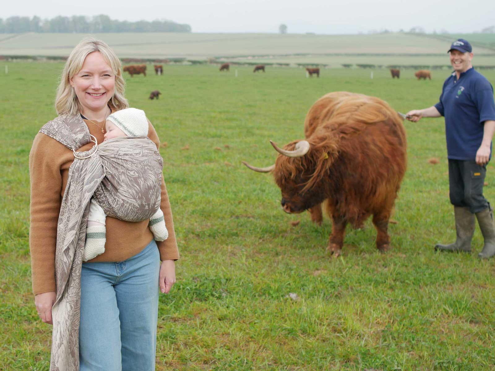 Woman holding a baby in a sling, standing next to a highland cow in a field with another person in the background.