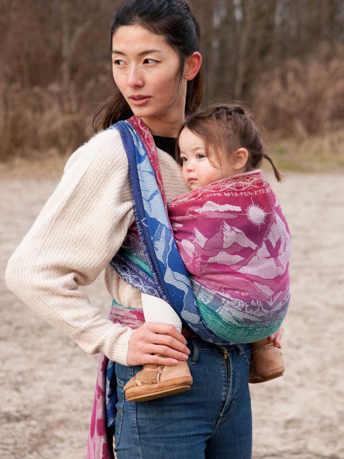 Woman carrying a child in a colourful baby carrier outdoors.