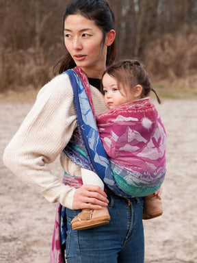 Woman carrying a child in a colourful baby carrier outdoors.