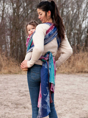 Woman carrying a child using a colourful baby wrap in an outdoor setting.