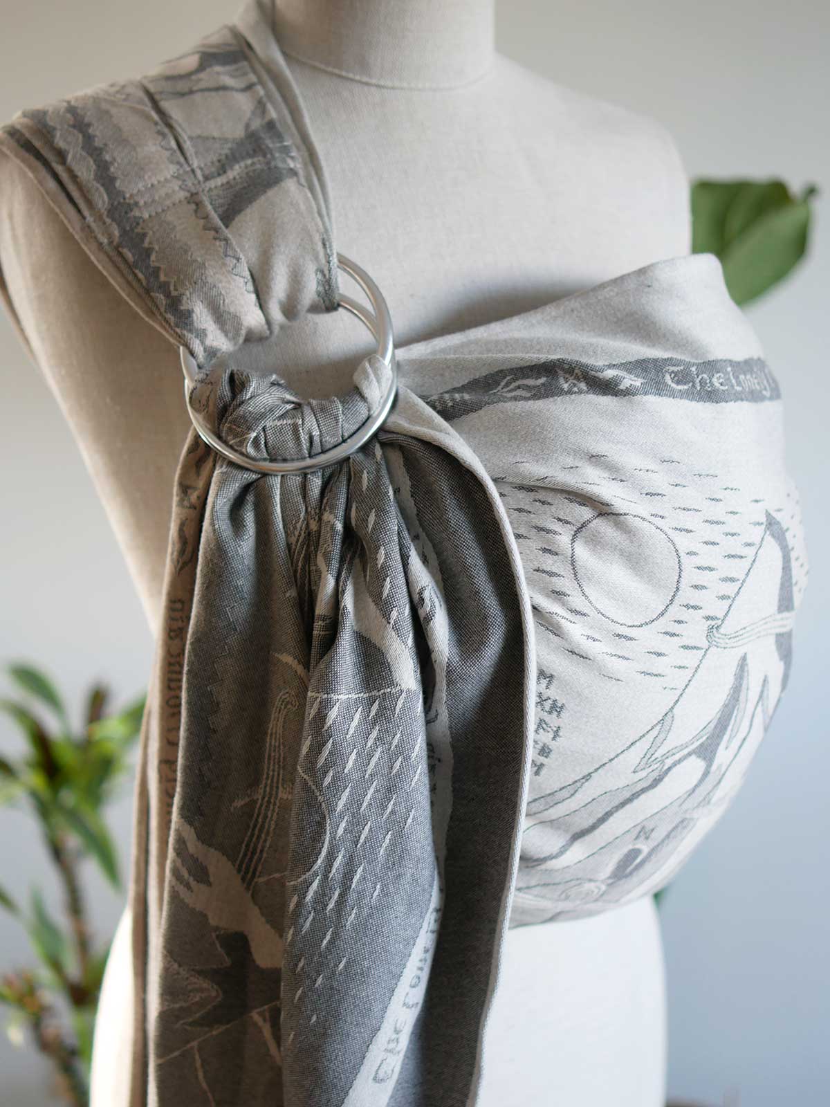 A baby ring sling in a gray and white print, shown on a mannequin torso with a silver metal ring. The fabric features a pattern of illustrations from Lord of the Rings.