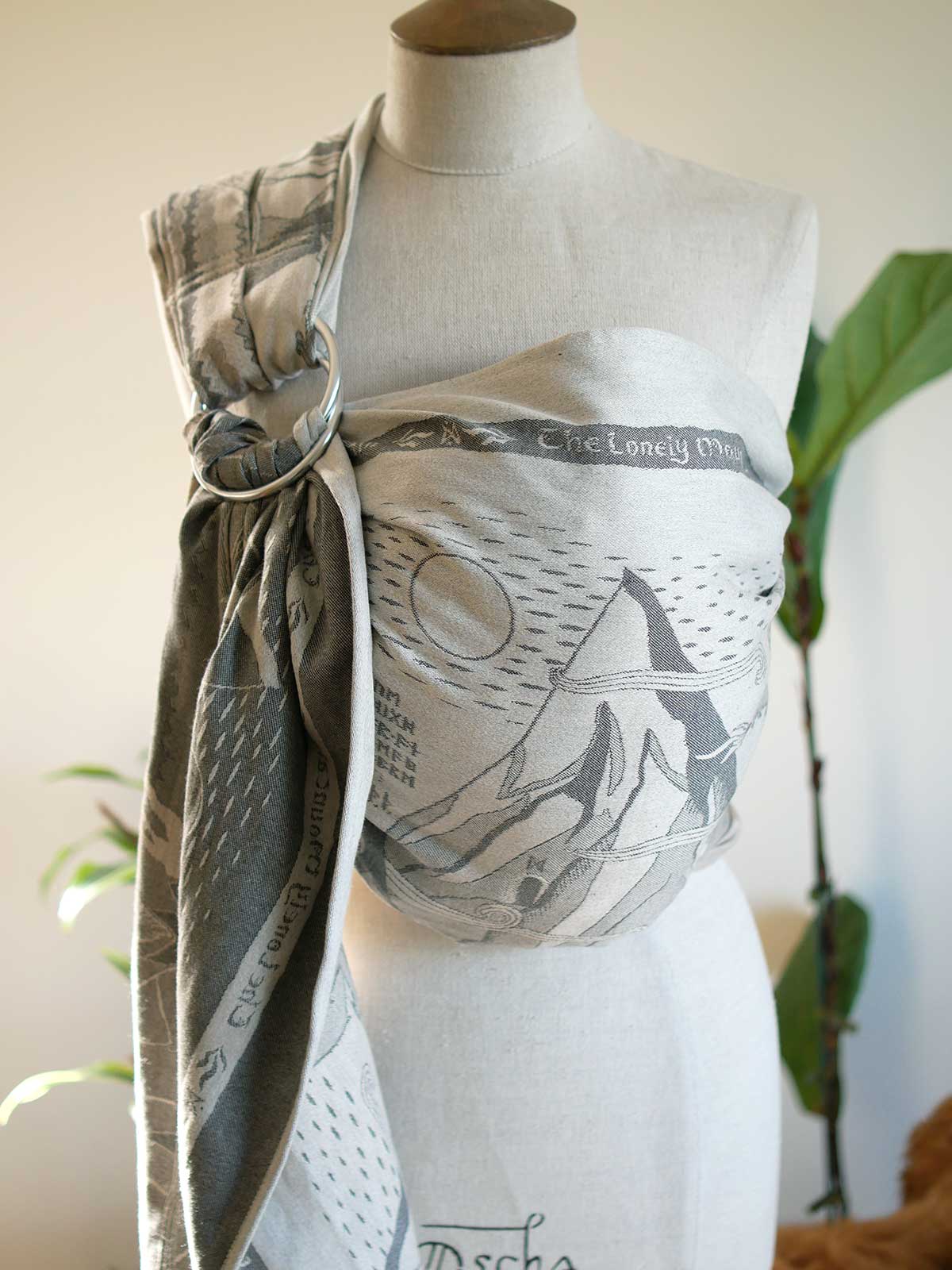 A baby ring sling with a gray and white graphic print, displayed on a beige fabric mannequin torso. The wrap features a mountain and moon design.