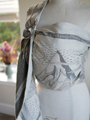A close-up of a fabric baby wrap or carrier draped on a white mannequin torso indoors. The wrap has a gray and off-white print with illustrated mountain shapes and text reading 'The Lonely Mountain.