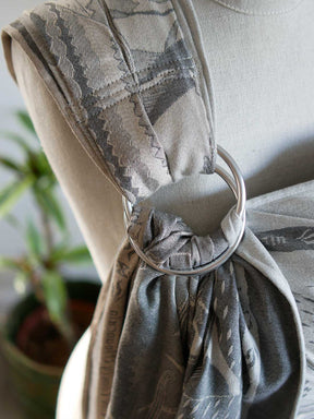A close-up of a baby ring sling with a gray and beige geometric pattern, secured with a large silver metal O-ring on a mannequin torso.