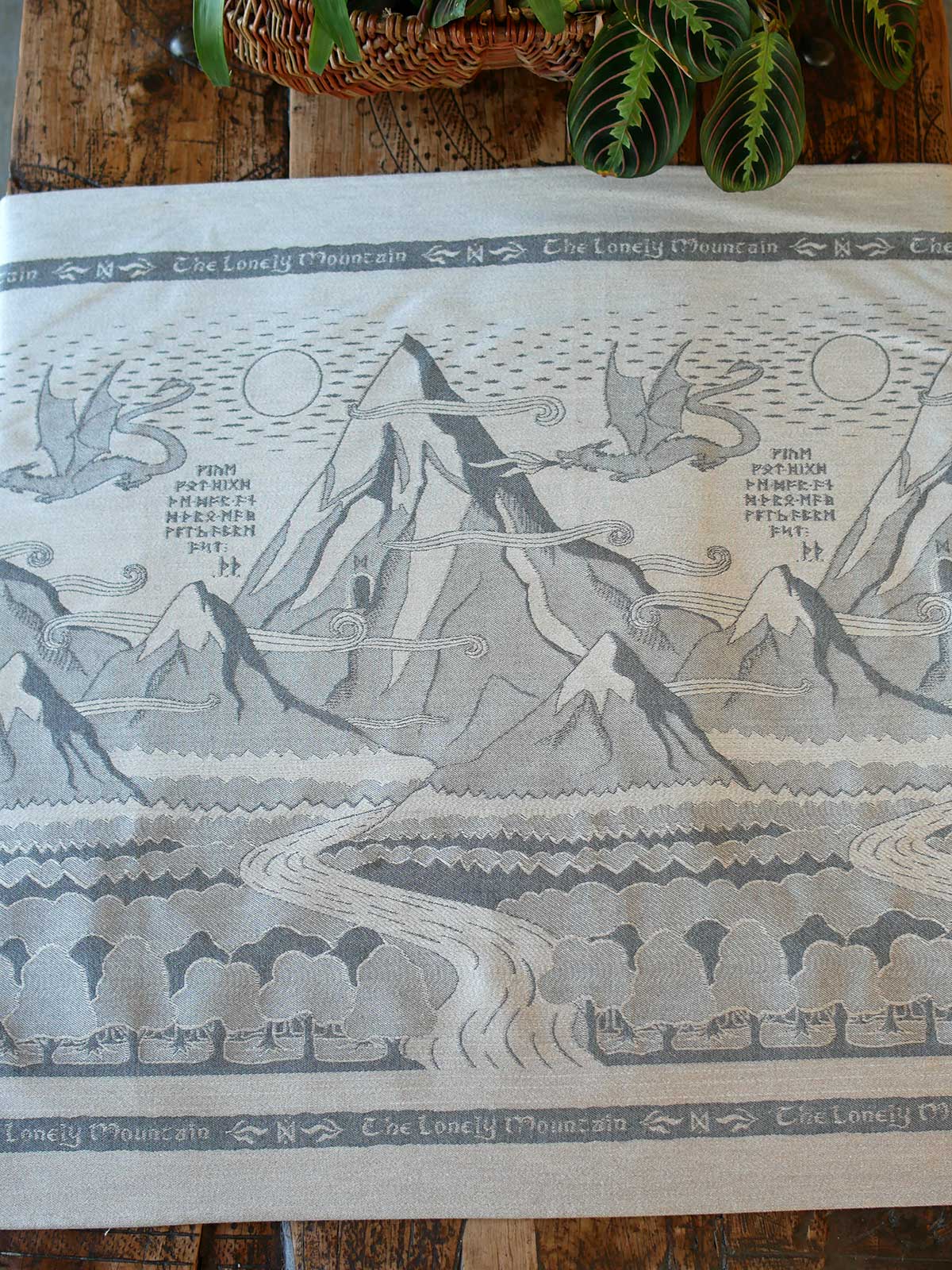 A light beige fabric featuring a detailed gray and black illustration of 'The Lonely Mountain' from Middle-earth, laid out on a rustic wooden table with green leaves at the top.