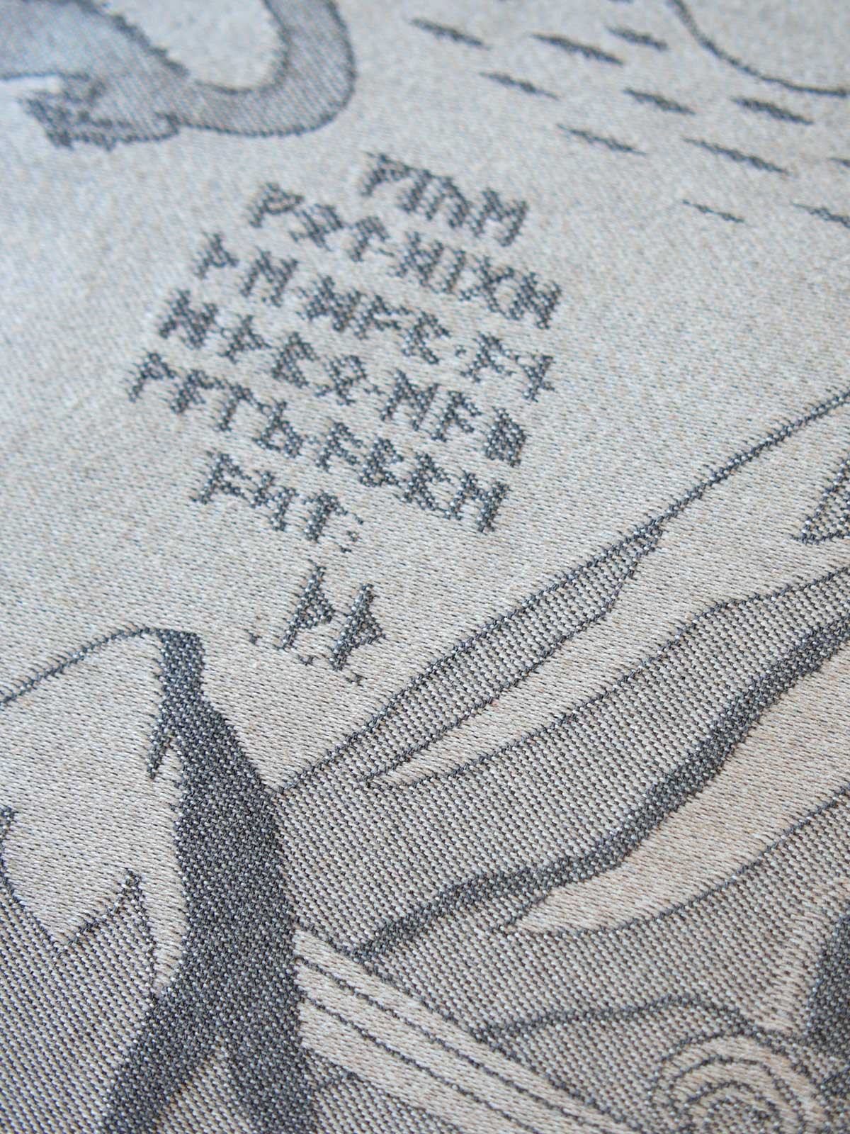 Close-up of a printed textile or fabric surface with gray line-art illustration and a cluster of dark gray characters on a light beige background.