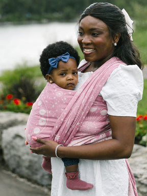Woman holding a child wrapped in a pink Lunae Isla Baby Wrap outdoors by a body of water.