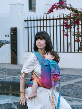 Woman carrying a child in a colorful sling outdoors near a fountain.