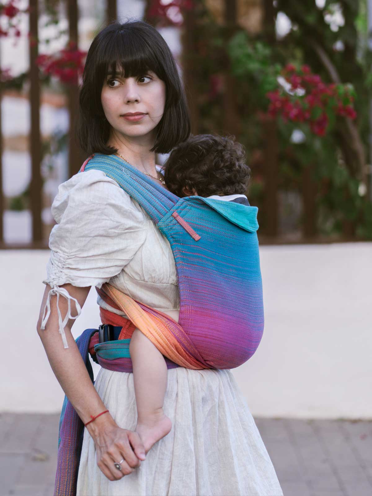 Woman holding a child in a colorful baby carrier with a blurred outdoor background. 