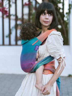 Model carrying a child in a teal, purple and orange baby carrier outdoors.