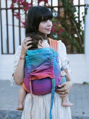 Model carrying a child in a Cairis baby carrier next to some magenta flowers.