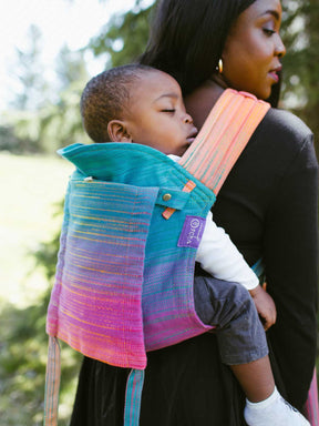Woman carrying a child on her back in a colorful Oscha Cairis baby carrier outdoors.