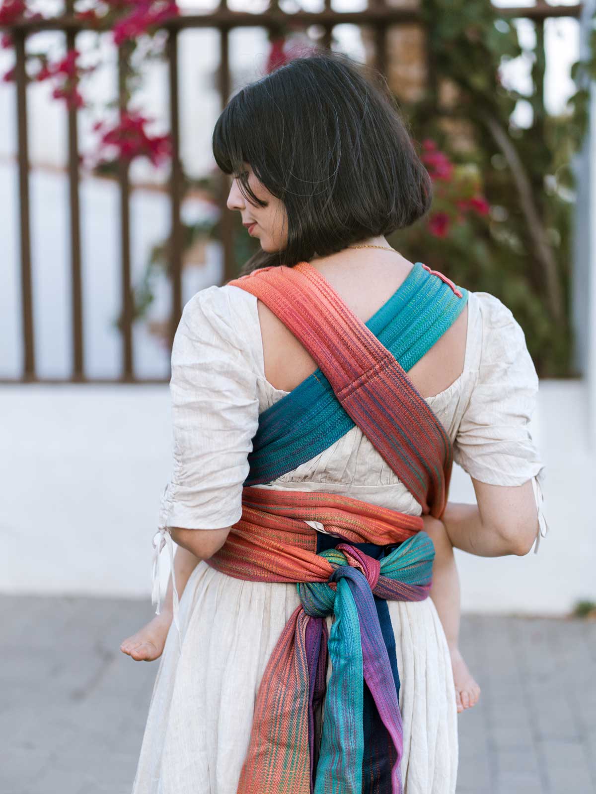 Back view of model using a baby carrier with crossed straps and knotted at the waist. 