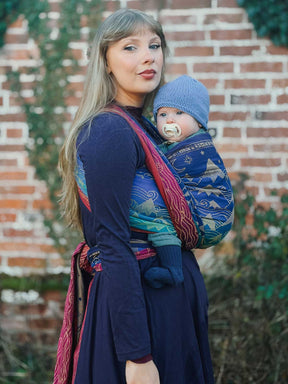 Woman carrying a baby in a sling against a brick wall.