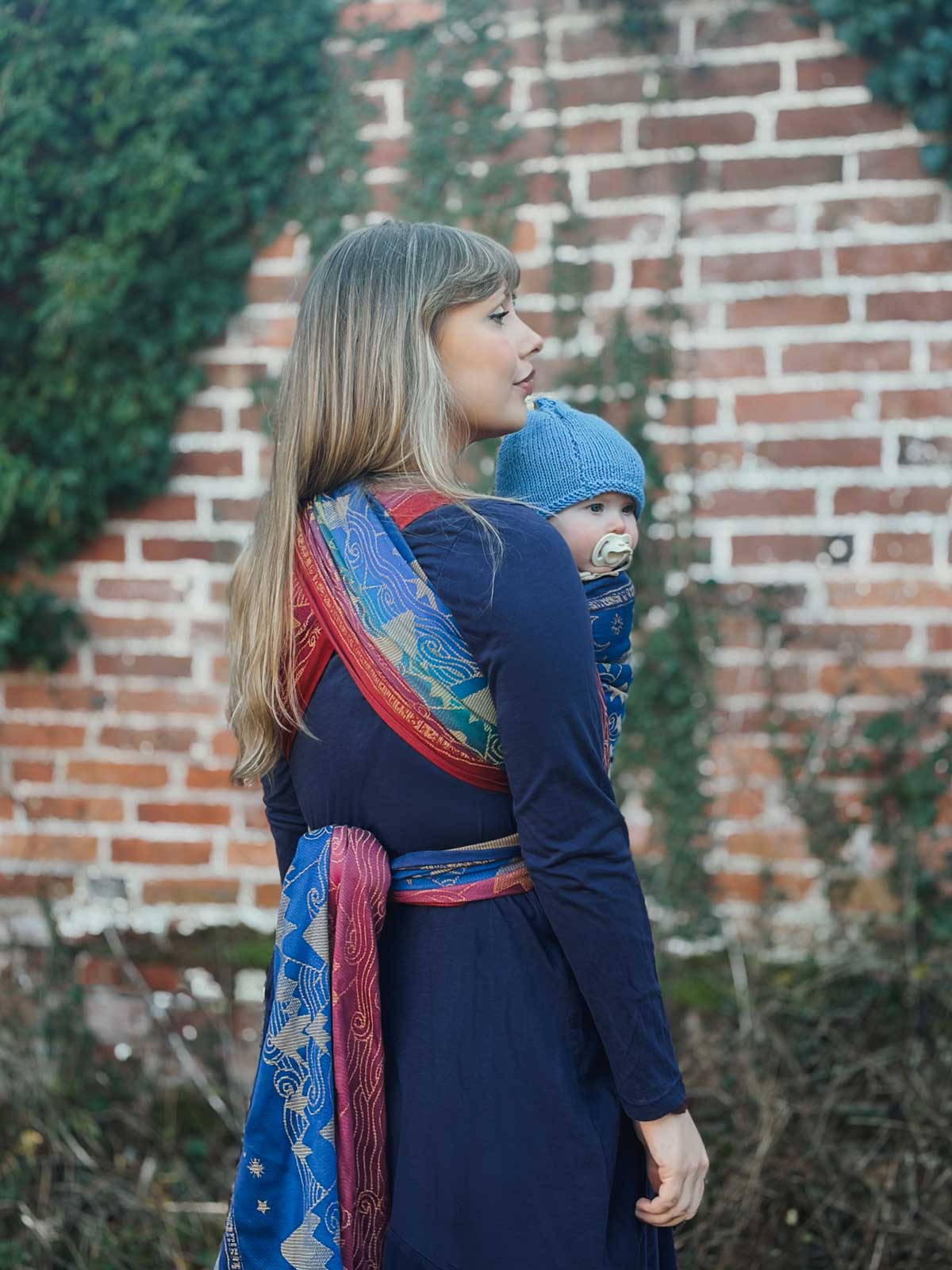 Woman in a blue dress carrying a child in a sling against a brick wall.