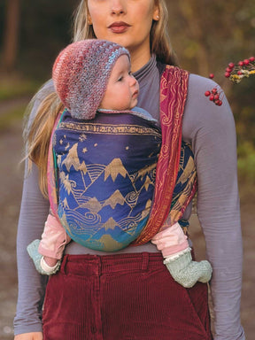 Woman carrying a child in a colorful baby carrier outdoors.