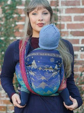 Woman holding a baby in a colorful woven wrap against a brick wall.