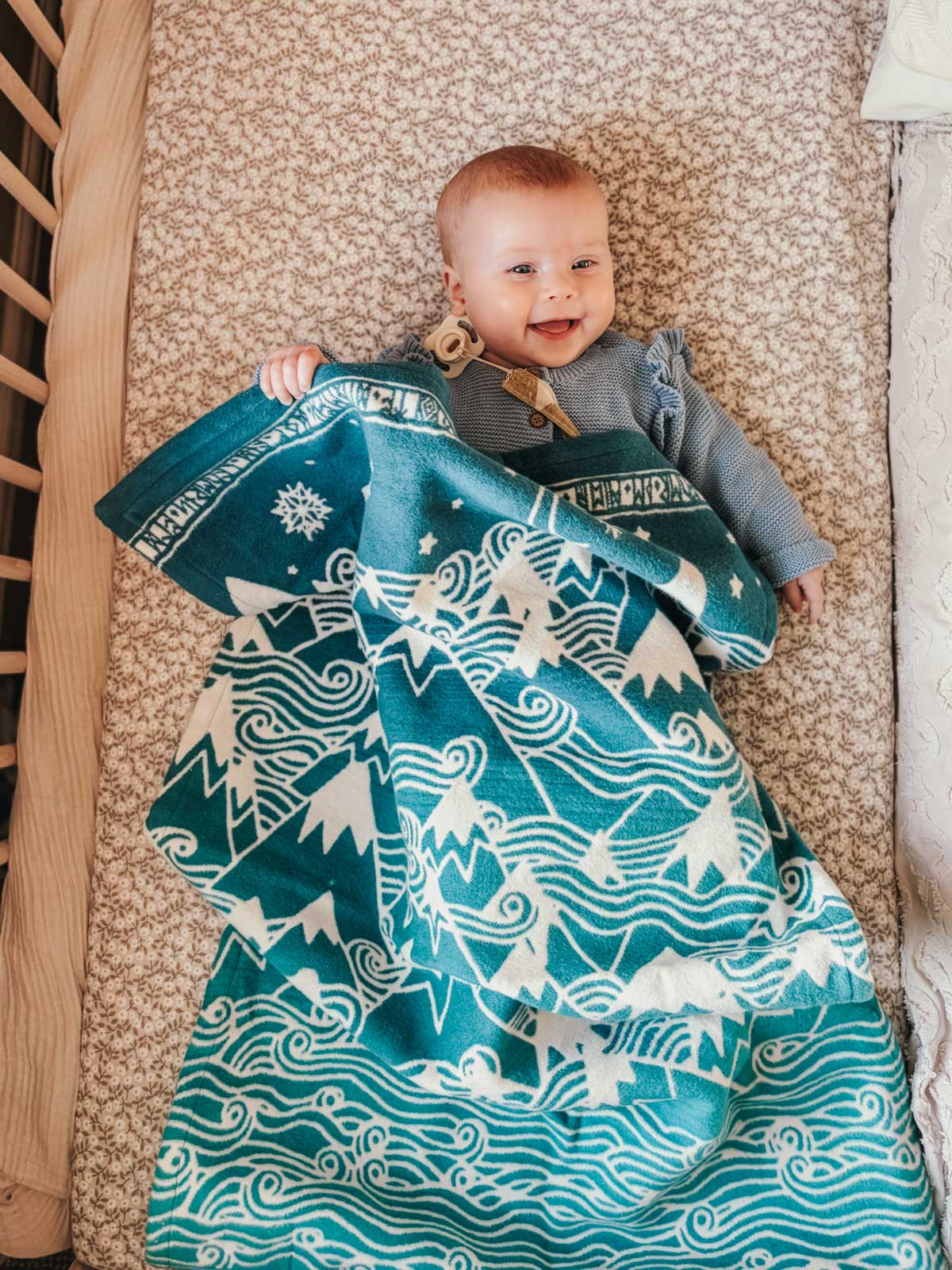 Baby lying in a cot with a teal blanket featuring Misty Mountains. 