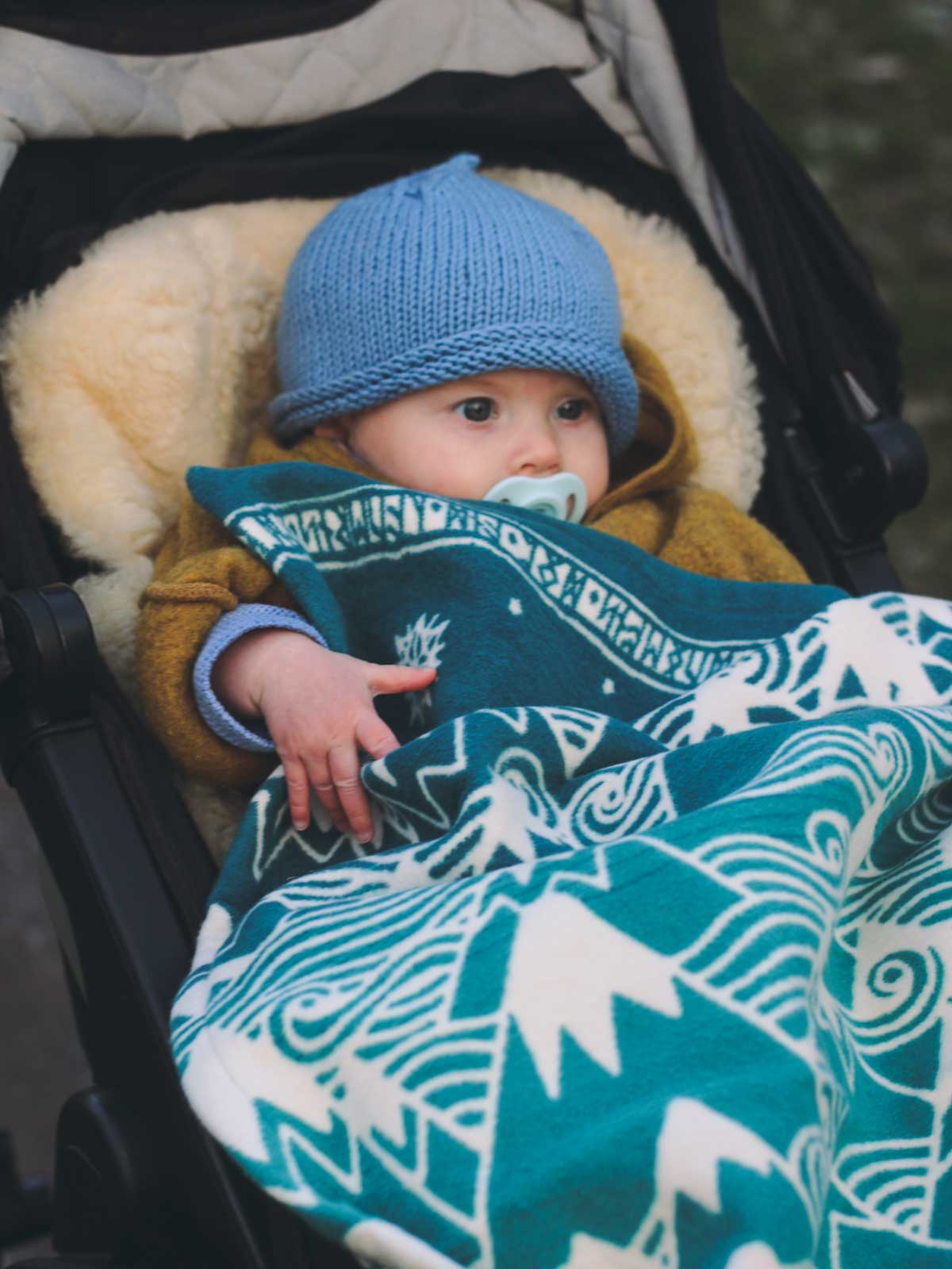 Baby in a pram wearing a blue knit hat and wrapped in a teal blanket with white Misty Mountains pattern.