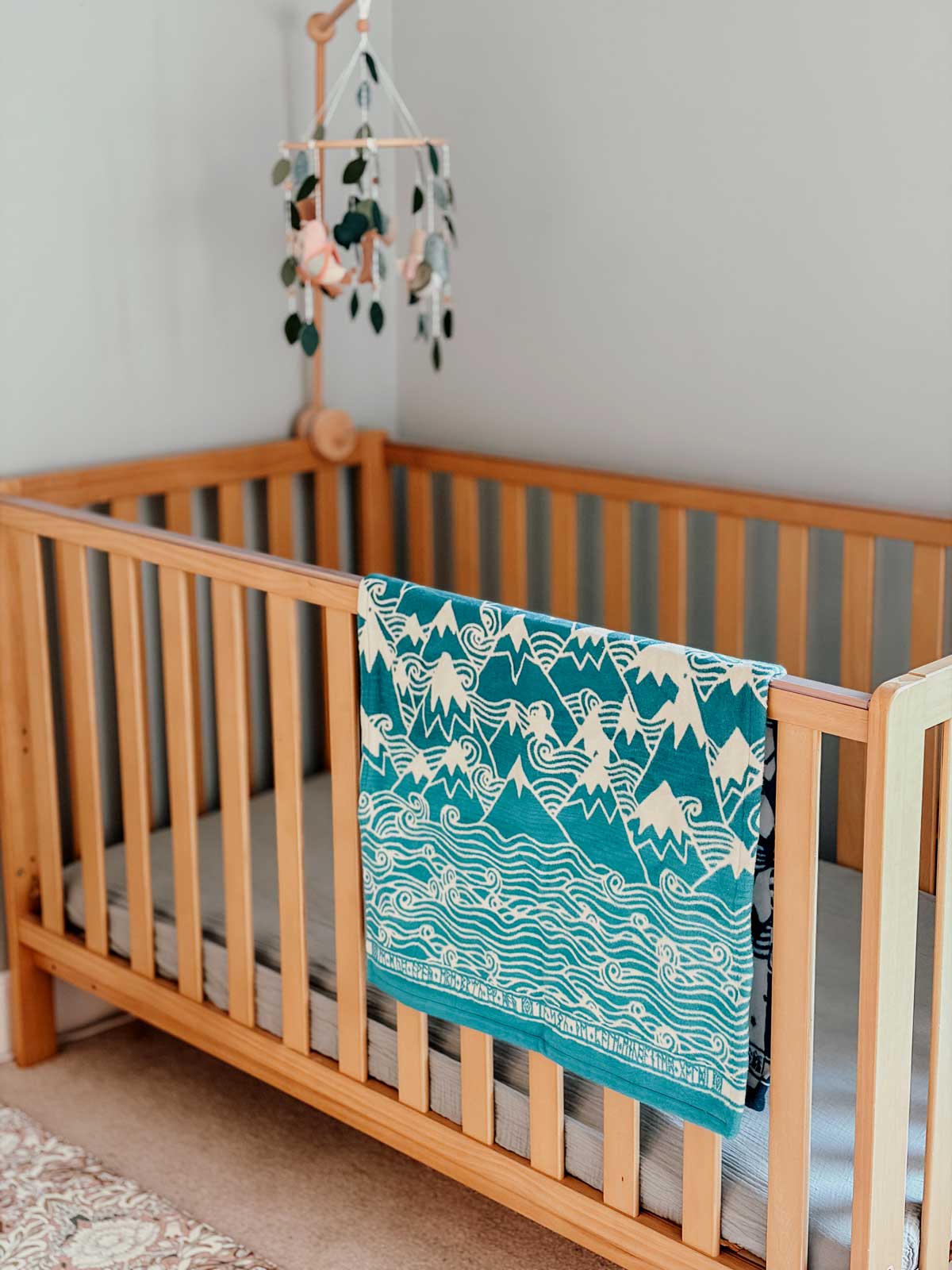 Wooden crib with a teal and white patterned blanket draped over the side, against a neutral wall.