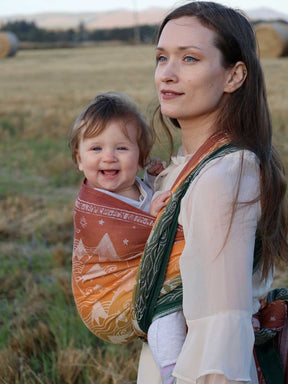 Woman holding a baby wrapped in a Misty Mountains Summit Ember Baby Wrap in a field.