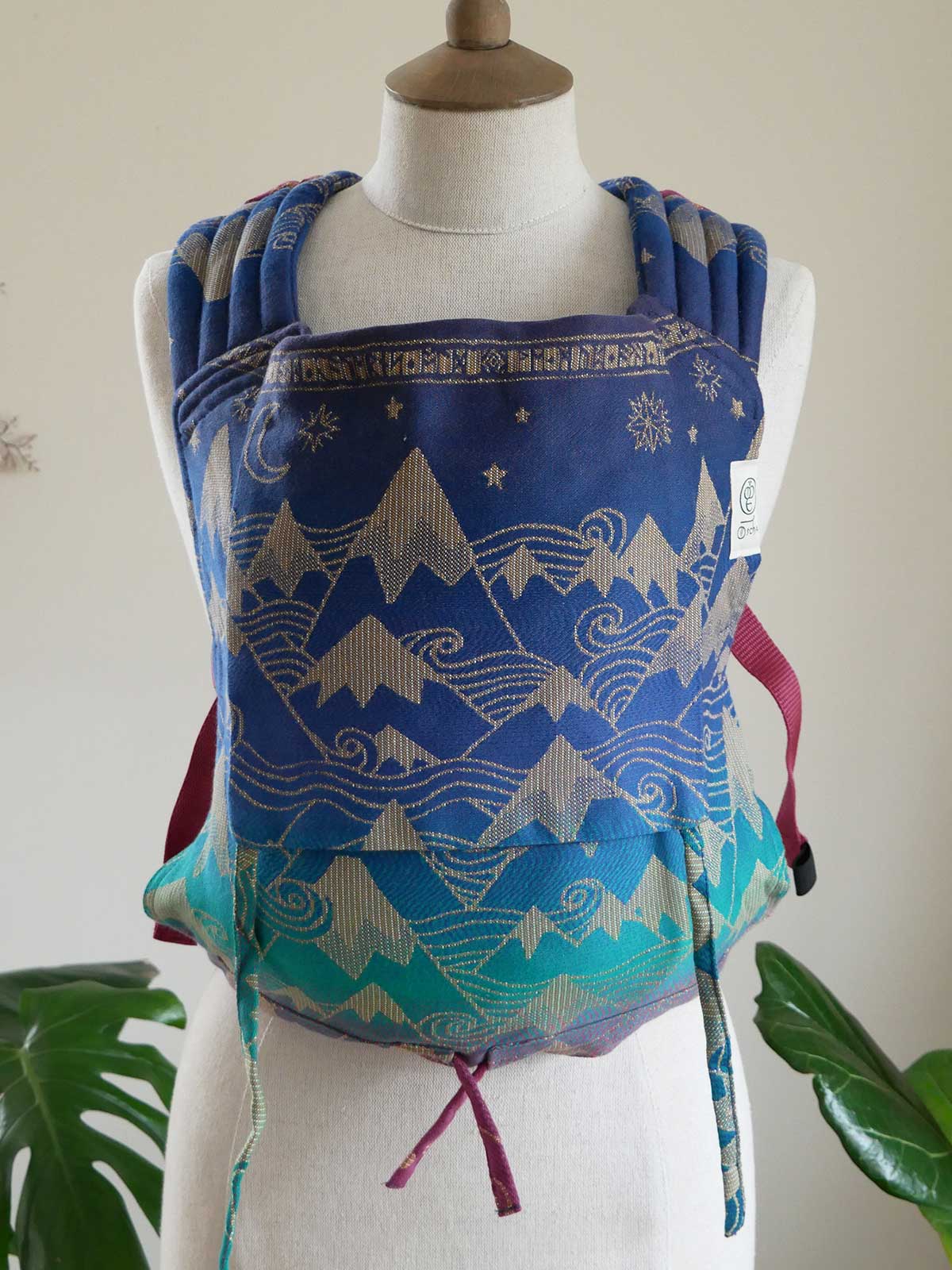 Blue and green baby carrier with mountain and wave patterns on a mannequin against a white background.