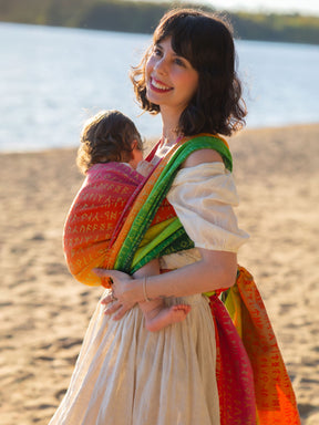 Woman holding a child wrapped in a colorful Moria Citrus Baby Wrap on a beach