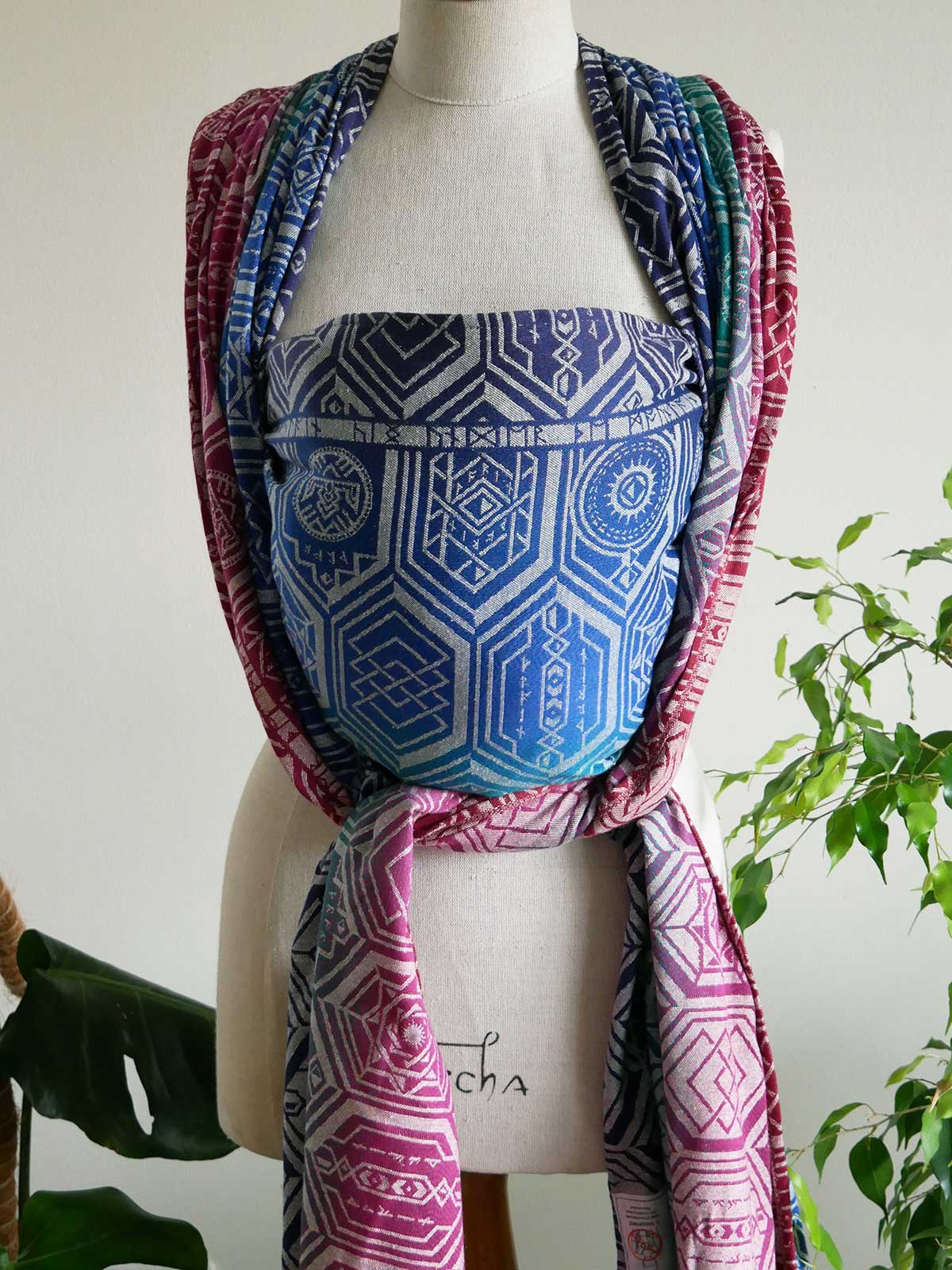 Oakenshield Loden baby wrap on a mannequin in jewel tones with a repeating shield pattern.
