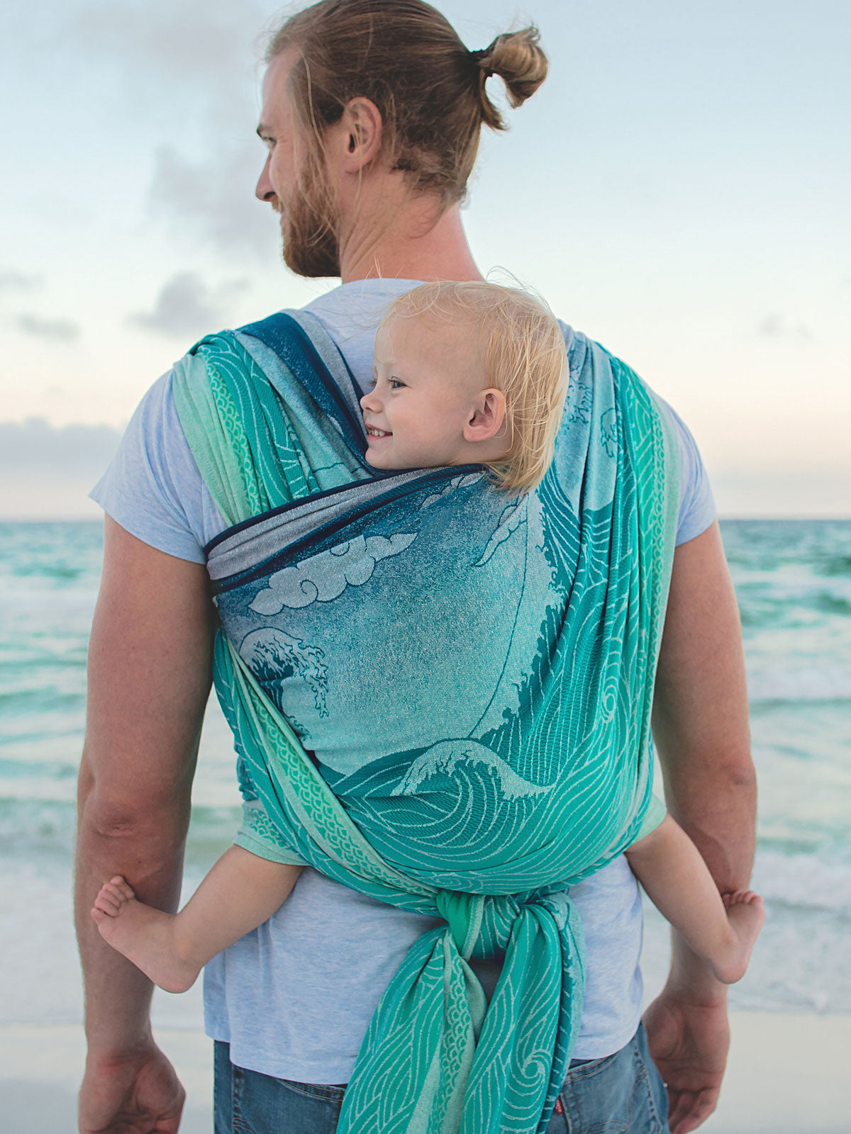 Man carrying a child on a beach using a teal Okinami Wipeout woven wrap.