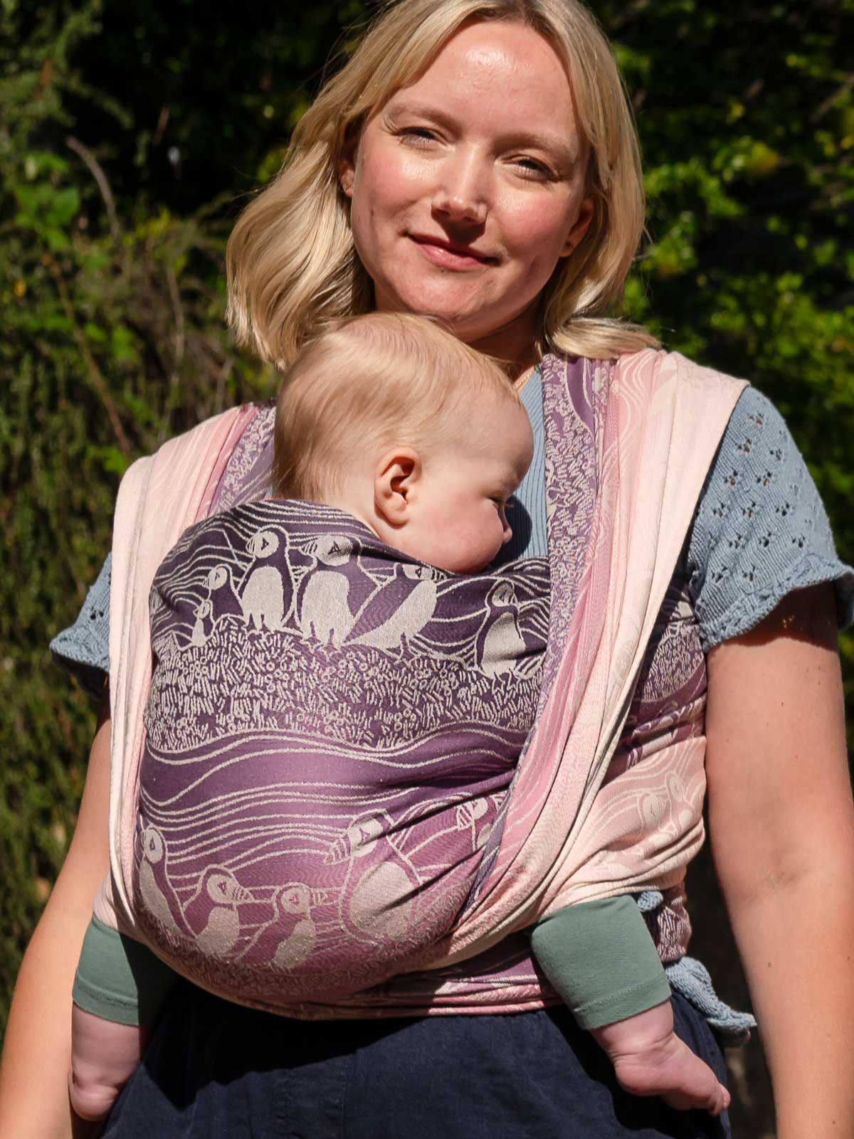 Front view of a woman wearing a baby in a pink and purple woveen wrap with puffin pattern