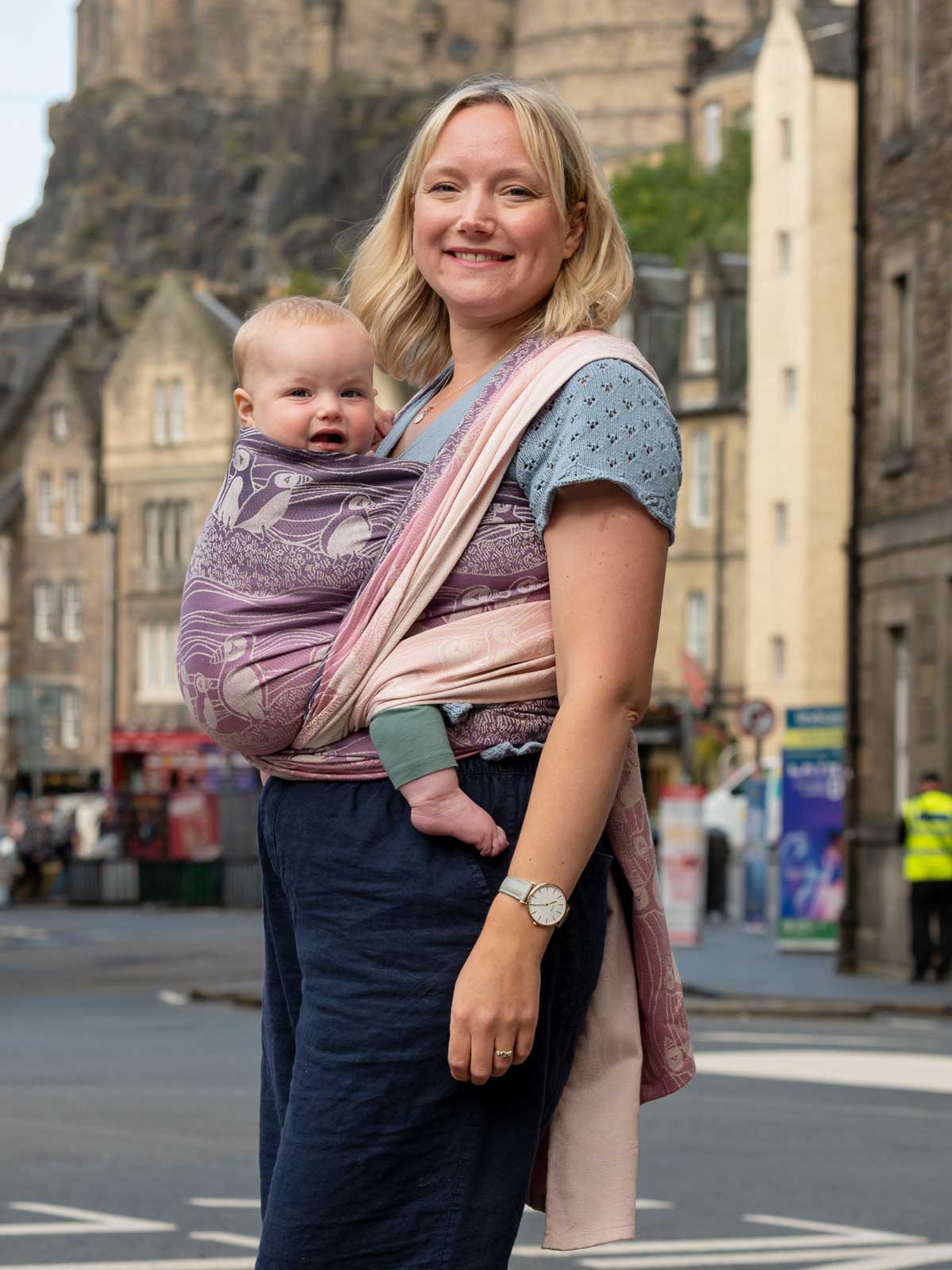 A woman with city scene in background wearing a baby in a pink and purple woven wrap with puffin pattern