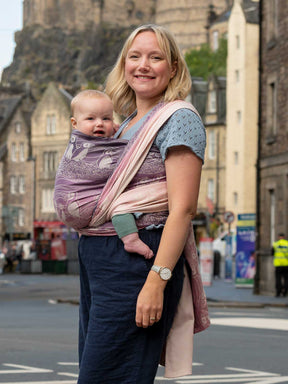 A woman with city scene in background wearing a baby in a pink and purple woven wrap with puffin pattern