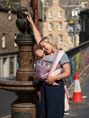 A woman wearing a baby in a pink and purple woveen wrap with puffin pattern touching a statue in Edinburgh