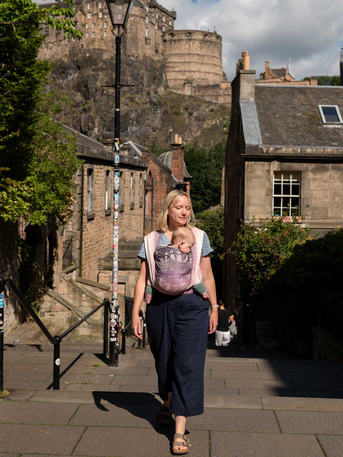 A woman wearing a baby in a pink and purple woveen wrap with puffin pattern with Edinburgh in the background