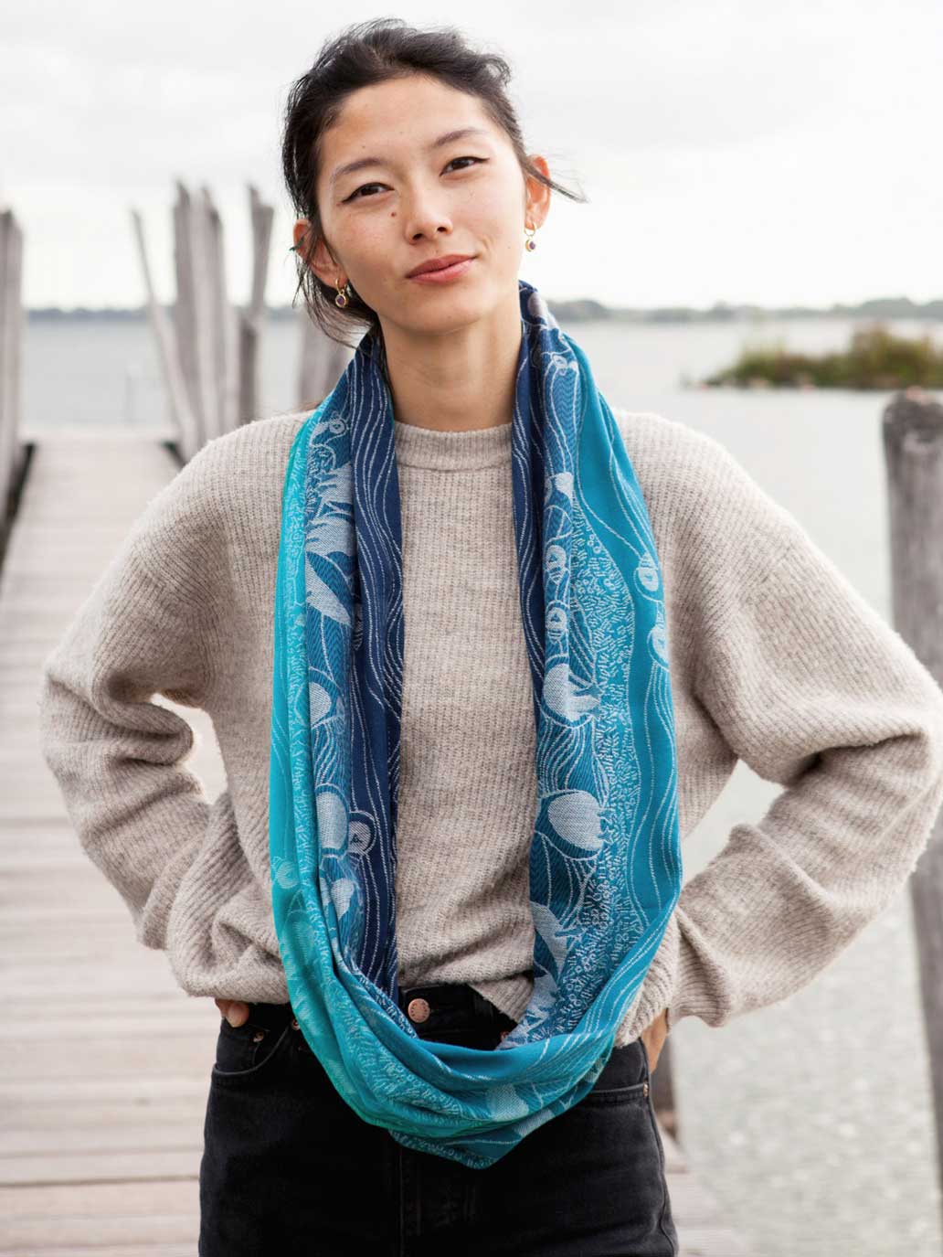 A woman standing on a pier wearing a teal and blue infinity loop with puffin pattern