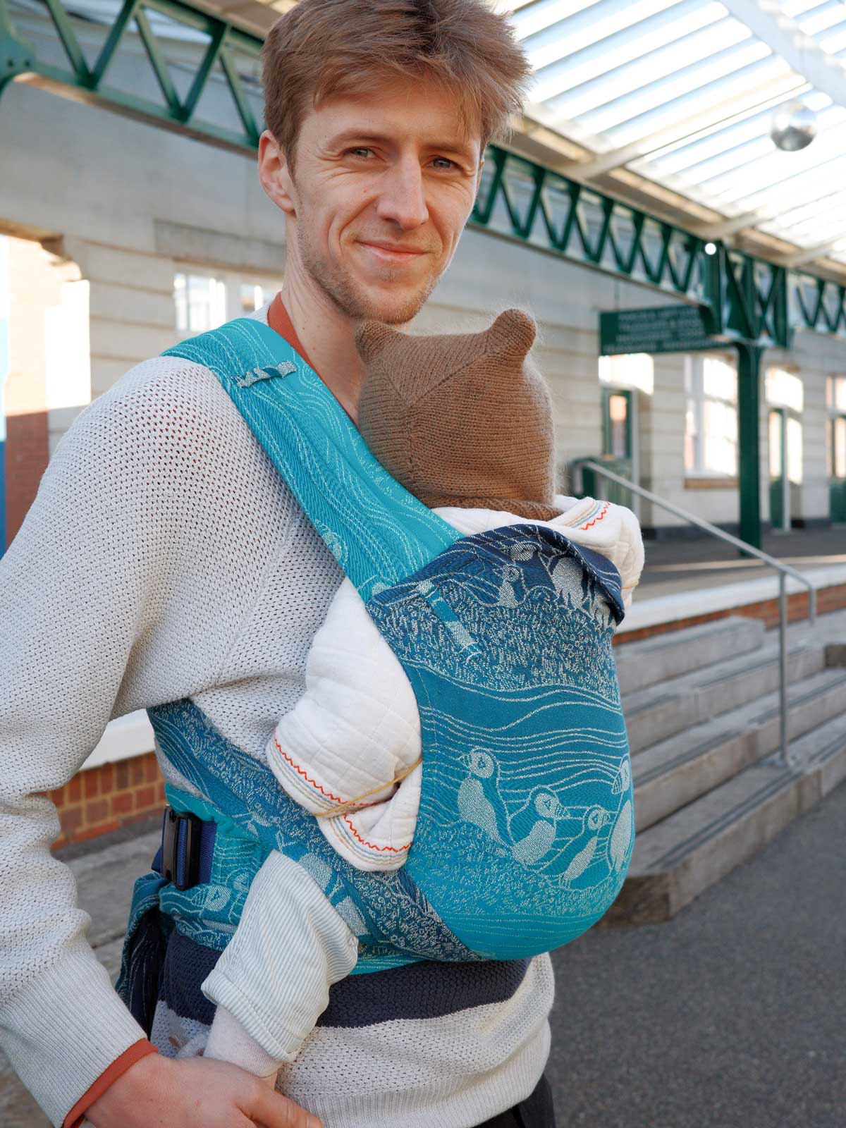 Person carrying a baby in a blue sling with Puffin design outdoors.